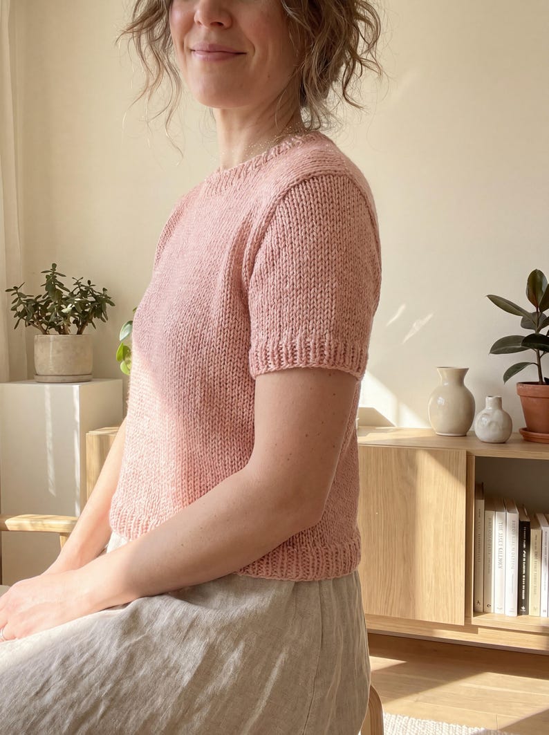 May include: A short-sleeved, crew-neck knit sweater in a soft, dusty pink color. The garment features a textured, hand-knit appearance with ribbed detailing along the neckline, sleeve cuffs, and the bottom hem, providing a structured fit.