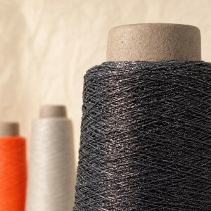 May include: A large cone of dark charcoal gray metallic thread with a visible shimmer, featuring a cardboard core. In the blurred background, two additional cones of thread are visible, one in a light gray and one in a bright orange color.