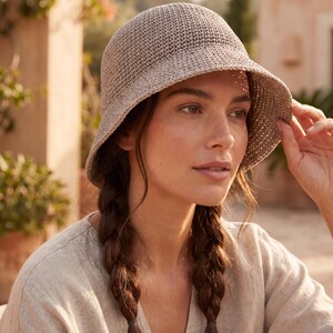 May include: A light beige crochet bucket hat with a rounded crown and a downward-sloping brim. The hat is constructed from a dense, textured yarn weave, featuring a consistent stitch pattern throughout the entire surface of the headwear.