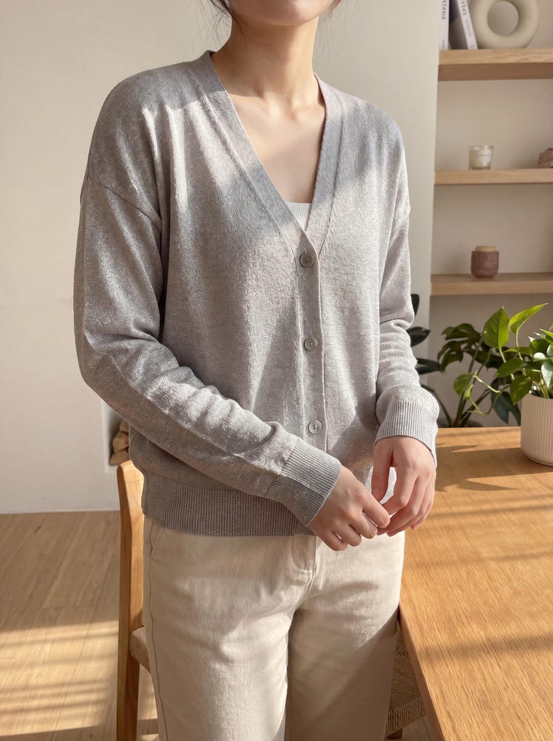 May include: A light gray long-sleeve cardigan with a V-neckline and a front button closure. The garment features ribbed cuffs and a ribbed hem, crafted from a fine-knit fabric. It is worn over a white top and paired with light beige trousers.