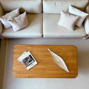 Op de afbeelding: Vogelperspectief van een woonkamer. Een lichtbeige bank met vier sierkussens staat boven een rechthoekige houten salontafel. Een tijdschrift en een blad-vormige decoratieve schaal liggen op de tafel.