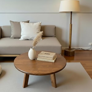 May include: A round, wooden coffee table with a natural finish. A small, white vase holds dried ornamental grass. Two books are stacked on the table. A beige sofa with pillows and a floor lamp are in the background.