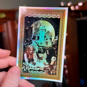 May include: A holographic sticker featuring a framed illustration of two women at a table, with a large moon and a house in the background. The image has a mystical theme, with a witchy aesthetic, and is surrounded by a gold border.