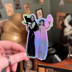 May include: A holographic sticker featuring two women, one holding a black cat and the other holding a white bird. The women are dressed in long gowns. The sticker has a colorful, iridescent finish.