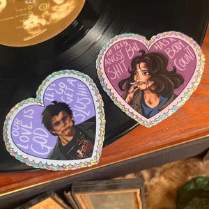 May include: Two heart-shaped stickers with glitter accents. The first sticker features a character with a bloody face and the text "Our love is God Let's go get a slushie". The second sticker features a character with a cigarette and the text "My teen angst bull shit has a body count".