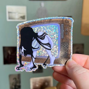 May include: A holographic sticker of a woman with long black hair crawling out of a vintage television set. The television is brown with a woodgrain finish and has a screen that is filled with glitter.