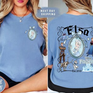 May include: Light blue t-shirt featuring a front and back design. The front has a portrait of Elsa. The back has the word "Elsa" and other Frozen characters. The shirt is a comfortable, casual style.