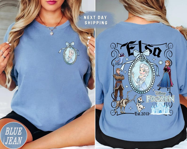 May include: Light blue t-shirt featuring a front and back design. The front has a portrait of Elsa. The back has the word "Elsa" and other Frozen characters. The shirt is a comfortable, casual style.