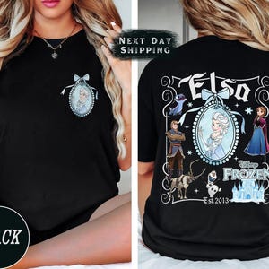 May include: Black t-shirt featuring a front pocket design of Elsa from Disney's Frozen. The back of the shirt displays a larger graphic with Elsa, other characters, and the text "Elsa" and "Disney Frozen Est. 2013".