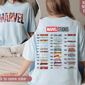 Puede incluir: Camiseta azul claro con la palabra "MARVEL" en un dise&ntilde;o de bloques multicolores en la parte delantera. La parte trasera de la camiseta muestra el logotipo de "MARVEL STUDIOS" y una lista de t&iacute;tulos de pel&iacute;culas y a&ntilde;os de lanzamiento.