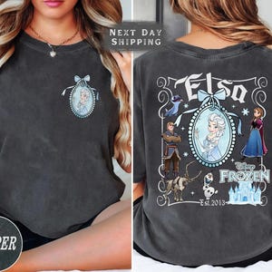 May include: Pepper-colored t-shirt featuring a front and back design. The front has a small oval portrait of Elsa. The back displays a larger design with Elsa, other characters, and the text "Elsa" and "Disney Frozen Est. 2013".