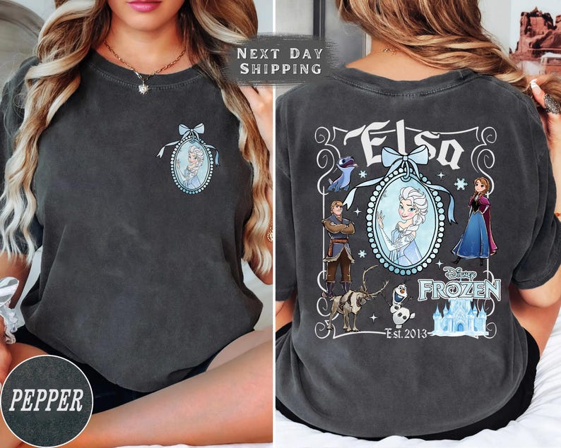 May include: Pepper-colored t-shirt featuring a front and back design. The front has a small oval portrait of Elsa. The back displays a larger design with Elsa, other characters, and the text "Elsa" and "Disney Frozen Est. 2013".