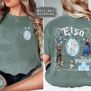 May include: Moss green t-shirt featuring a front chest graphic of Elsa in a blue oval frame with a bow. The back of the shirt displays the word "Elsa" and characters from the Disney movie Frozen, including Anna, Kristoff, and Olaf.