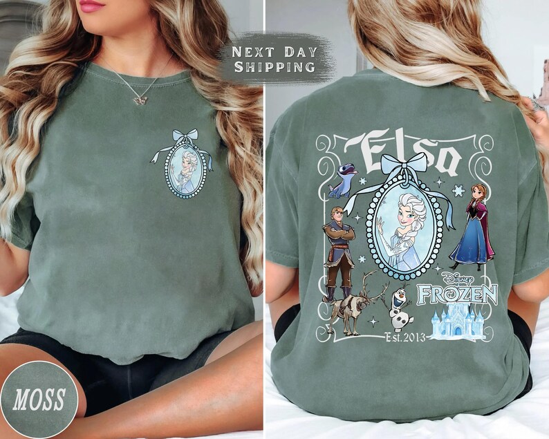 May include: Moss green t-shirt featuring a front chest graphic of Elsa in a blue oval frame with a bow. The back of the shirt displays the word "Elsa" and characters from the Disney movie Frozen, including Anna, Kristoff, and Olaf.