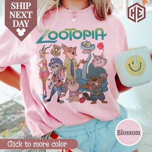 Puede incluir: Una camiseta rosa claro de cuello redondo con la palabra "Zootopia" en verde y un gr&aacute;fico de personajes de la pel&iacute;cula animada. La camiseta tambi&eacute;n tiene el texto "Click to more color" y una muestra de color "Blossom".