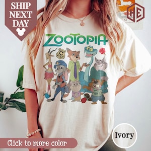 Puede incluir: Camiseta de color marfil con la palabra "ZOOTOPIA" en verde y personajes de dibujos animados de la pel&iacute;cula. La camiseta tiene cuello redondo y mangas cortas. El texto "Click to more color" est&aacute; en la parte inferior.