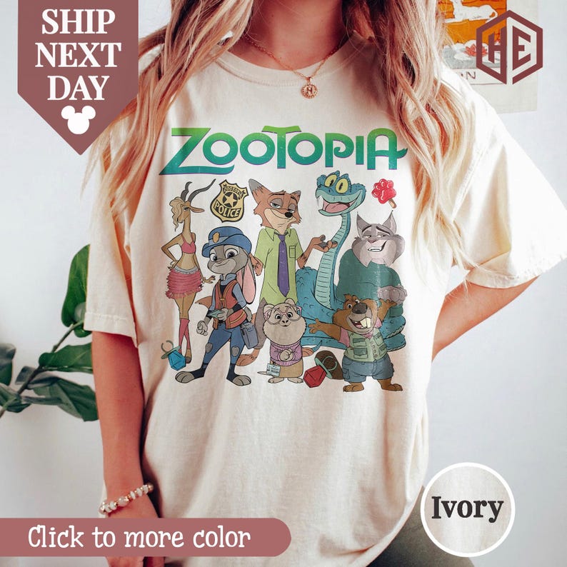 Puede incluir: Camiseta de color marfil con la palabra "ZOOTOPIA" en verde y personajes de dibujos animados de la pel&iacute;cula. La camiseta tiene cuello redondo y mangas cortas. El texto "Click to more color" est&aacute; en la parte inferior.