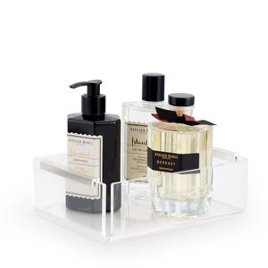 May include: A clear acrylic tray holding a black liquid soap dispenser, two perfume bottles, and a clear bottle with a black cap. The bottles have black labels with white text. The tray is on a white background.