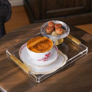 May include: A cup of coffee with a floral design sits on a white saucer, placed on a decorative doily. The cup and saucer are on a clear acrylic tray with gold handles. A small glass dish with chocolates is also on the tray.