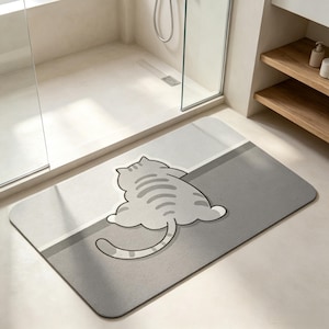 May include: A rectangular bath mat featuring a light gray and dark gray color-blocked design. A stylized illustration of a plump cat with dark gray stripes is centered on the mat, depicted from behind with its long tail curved to the left side.