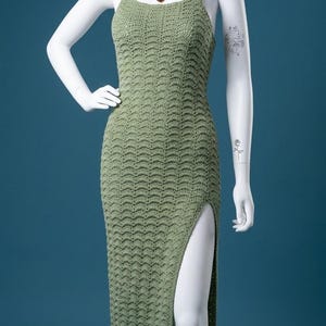 May include: A sage green crochet dress with a high side slit, displayed on a mannequin. The dress has a textured, wave-like pattern and spaghetti straps. A heart-shaped pendant necklace is visible.