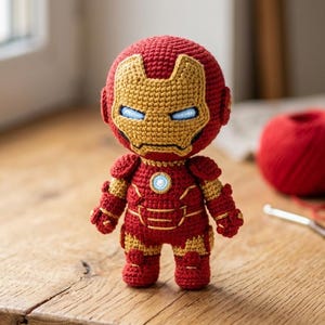 Puede incluir: Muñeco de Iron Man tejido a mano en rojo y dorado. El muñeco presenta un casco detallado con ojos azules y un emblema en el pecho. Una madeja de lana roja, un ganchillo y tela están en el fondo. El muñeco mide aproximadamente 15 cm de alto.