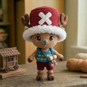 以下が含まれることがあります： 白い十字の赤い帽子、茶色の角、白い毛皮のトリムが付いたキャラクターの手作りのかぎ針編み人形。人形は青いベスト、赤いショーツ、小さなバッグを着ています。他の編み人形と糸が背景にあります。