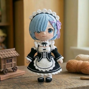 Könnte beinhalten: Handgefertigte Häkelpuppe mit blauen Haaren und einem schwarz-weißen Dienstmädchen-Outfit. Die Puppe hat große blaue Augen und eine kleine lila Schleife. Ein Miniatur-Holzhaus auf Rädern befindet sich in der Nähe, zusammen mit Garn und Holzspulen.