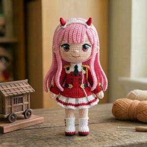Pode incluir: Boneca de crochê feita à mão com cabelo rosa e chifres vermelhos, vestindo uma roupa vermelha e branca. A boneca está em uma superfície de madeira. Outros itens de crochê e uma casa de madeira em miniatura estão no fundo.