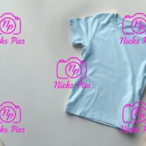 May include: A light blue t-shirt is displayed on a neutral background, surrounded by draped fabric in shades of cream and white. The soft fabric creates a gentle contrast with the smooth texture of the shirt, offering a minimalist aesthetic.