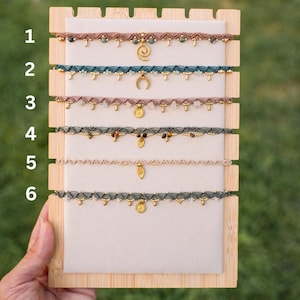 Pode incluir: Seis gargantilhas de macramé exibidas num suporte de madeira clara, numeradas de um a seis. Cada colar apresenta um cordão entrançado de várias cores, pequenas contas metálicas e um pendente único em tom dourado com formas variadas.