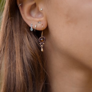 Könnte beinhalten: Nahaufnahme eines Ohres mit mehreren Piercings und einem hängenden Ohrring. Der Ohrring hat einen braunen Edelstein, Makramee-Details und einen goldfarbenen Metallakzent. Weitere Piercings umfassen silber- und goldfarbene Ohrstecker.