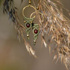 May include: Close-up of handmade earrings with a bohemian style. The earrings feature gold-coloured hoops and hooks, with olive green macrame detailing and small, round, red beads. The earrings are hanging in front of a blurred background of dried grass.