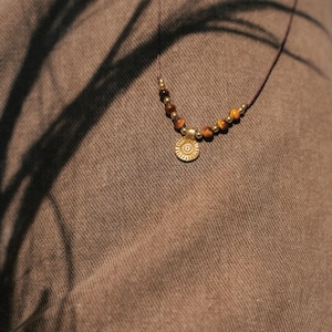 Puede incluir: Un collar con un cordón fino de color marrón oscuro adornado con pequeñas cuentas redondas de ojo de tigre y espaciadores metálicos dorados. Un colgante circular dorado con un patrón de rayos de sol y un punto central cuelga del centro.