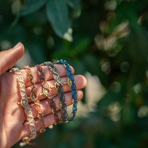 Könnte beinhalten: Fünf Makramee-Armbänder in Beige, Braun, Grau und Blau werden gezeigt. Jedes Armband hat ein zentrales dekoratives Element und kleine goldene Perlen. Der Hintergrund ist verschwommenes grünes Laub. Gehalten in einer Hand.