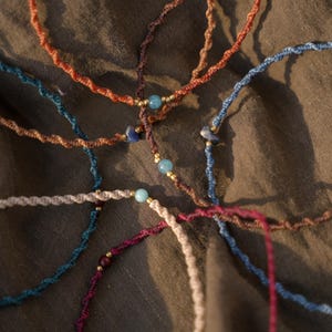 Pode incluir: Uma coleção de colares de macramé feitos à mão em várias cores, incluindo turquesa, laranja e bordô. Cada colar apresenta pequenas contas e um design de cordão torcido, sobre um fundo de tecido verde escuro.