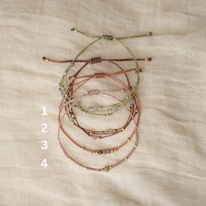 Könnte beinhalten: Vier verstellbare Kordelarmbänder, beschriftet mit 1 bis 4. Jedes Armband besteht aus einer dünnen farbigen Schnur, kleinen Metallperlen und zentralen Steinakzenten. Die Armbänder sind vertikal angeordnet und zeigen verschiedene Erdtöne.