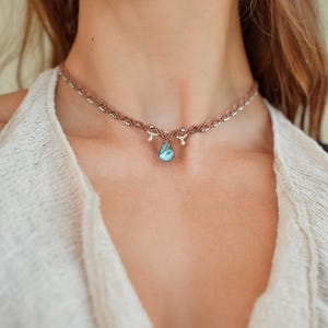 Op de afbeelding: Een bruine macramé choker ketting met een centrale druppelvormige labradoriet edelsteen hanger met iriserende blauwe flitsen, geaccentueerd door kleine zilverkleurige metalen kralen geweven door het ingewikkelde koordontwerp.