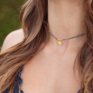 Può includere: Una collana girocollo con un cordino sottile e testurizzato color verde salvia e un piccolo ciondolo circolare dorato con un motivo a raggiera. Il ciondolo è centrato e affiancato da due minuscole perline dorate su entrambi i lati.