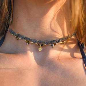 Può includere: Una collana girocollo in macramè grigio con piccole perline color oro e accenti in pietra occhio di tigre marrone. Il design include un ciondolo centrale color oro a forma di foglia appeso al cordino intrecciato, con piccole gocce metalliche.
