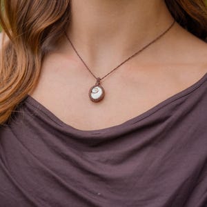 Könnte beinhalten: Eine Halskette mit einem kleinen, ovalen weißen Muschelanhänger mit natürlichem Spiralmuster. Die Muschel ist in eine dunkelbraune Makramee-Schnurfassung eingefasst, die als dünne, geflochtene Kordel am Schlüsselbein entlang verläuft.