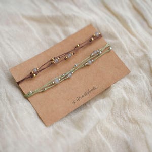 Può includere: Due braccialetti in cordino sottile presentati su un cartoncino rettangolare marrone. Un braccialetto è marrone scuro con perline dorate e piccole pietre grigie, l'altro è verde chiaro con perline argentate e pietre grigie. È stampato il testo ig: @earthyknots_.