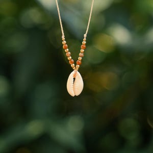 Könnte beinhalten: Eine zarte Halskette mit einem Anhänger aus natürlicher Muschel. Die Halskette hat eine beige Kordel mit goldfarbenen Perlen und braunen Edelsteinperlen. Die Muschel hat eine cremeweiße Farbe. Der Hintergrund ist ein verschwommenes Grün.