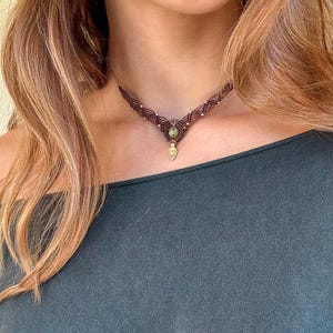 Pode incluir: Um colar gargantilha de macramé castanho com uma conta de pedra verde redonda ao centro e um pequeno pendente dourado em forma de gota. O cordão é atado num padrão geométrico repetitivo com pequenos detalhes metálicos incorporados.