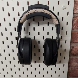 May include: Black over-ear headphones with a tan and black stitched headband are mounted on a white pegboard. The pegboard is against a brick wall. The headphones are designed for listening to music or other audio.