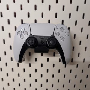 May include: A white and black PlayStation 5 DualSense wireless controller. The controller features a central touchpad, directional buttons, and action buttons. The controller is mounted on a white pegboard.