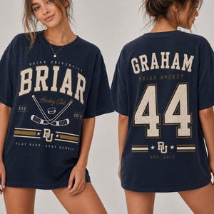 Könnte beinhalten: Ein marineblaues Kurzarm-T-Shirt mit einem abgenutzten cremefarbenen und goldenen Aufdruck. Die Vorderseite zeigt 'BRIAR UNIVERSITY' mit gekreuzten Hockeyschlägern und 'EST. 2015'. Die Rückseite zeigt 'GRAHAM', die Nummer '44' und 'BRIAR HOCKEY'.
