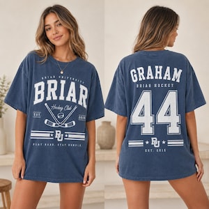 May include: A blue short-sleeve t-shirt featuring white graphics. The front displays 'BRIAR UNIVERSITY', 'Hockey Club', 'EST. 2015', and 'PLAY HARD, STAY HUMBLE' with hockey sticks. The back shows 'GRAHAM', 'BRIAR HOCKEY', and the number '44'.