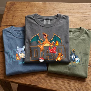Könnte beinhalten: Drei gefaltete T-Shirts in Blau, Grau und Grün, jedes mit einem anderen Pokemon-Motiv. Das graue Shirt hat ein großes "DAD"-Design mit einer Charizard-Illustration. Das blaue Shirt hat ein "M"-Design mit einem Schiggy und Glutexo. Das grüne Shirt hat ein "D"-Design mit einem Plinfa.