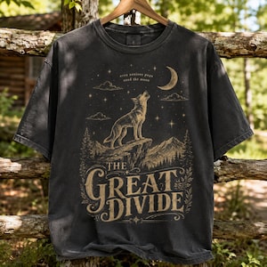 May include: A charcoal grey short-sleeve t-shirt featuring a tan graphic of a wolf howling at a crescent moon on a cliff. The design includes stars, clouds, pine trees, mountains, and the text "even anxious pups need the moon" and "THE GREAT DIVIDE".